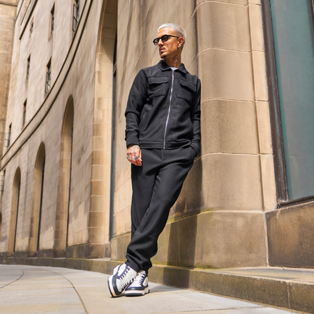 A man wearing a black zip-up jacket and matching black sweatpants stands on a stone sidewalk next to a beige stone building, wearing black and white sneakers.